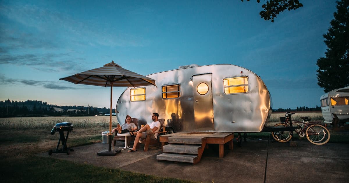 Silver Airstream-style caravan at dusk with a couple relaxing outside under an umbrella at a scenic campsite